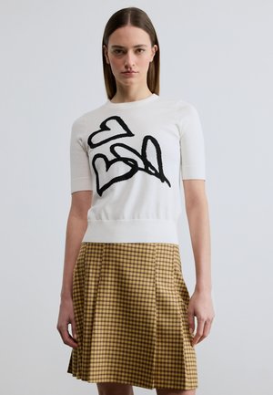 Mladá žena s rovnými hnědými vlasy má na sobě bílé tričko s krátkými rukávy s černými srdíčky a žlutou plisovanou sukní s kostkovaným vzorem, stojí před bílým pozadím.