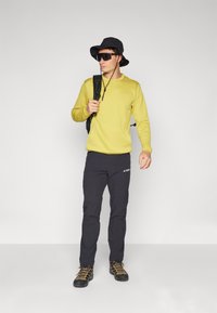 Cămașă galbenă cu mânecă lungă, pantaloni de drumeție negri cu logo, pălărie neagră tip bucket, ochelari de soare și bocanci de drumeție maronii cu șirete galbene.