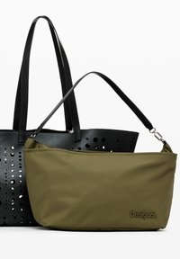 Sac fourre-tout noir perforé avec de longues anses à côté d'une pochette olive verte zippée arborant un logo subtil. Les deux sacs ont des textures lisses.