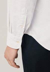 Camisa blanca de manga larga con puños abotonados, tejido texturizado, dobladillo recto y pantalones oscuros en contraste, que enfatizan las líneas limpias y la simplicidad.