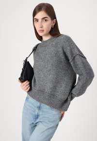 Šedý pletený svetr s žebrovaným lemem a manžetami. V kombinaci se světle modrými džíny a malou černou koženou kabelkou. Jednoduchý design, měkká textura.