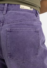 Jeans in denim viola con finitura opaca, caratterizzati da cuciture a contrasto e una sola tasca posteriore. L'etichetta vicino alla vita riporta "EST. 1992."