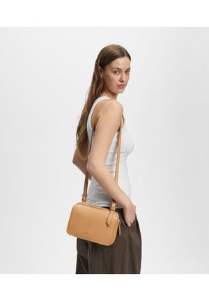 Frau mit langen Haaren, die ein weißes Tanktop und dunkle Hosen trägt, mit einer beigen Leder-Umhängetasche über der Schulter.