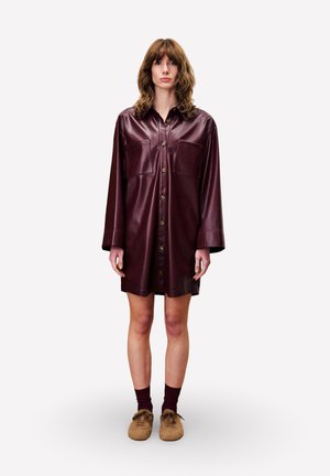 Femme aux cheveux bruns ondulés portant une robe chemise en cuir bordeaux à boutons, des chaussettes bordeaux et des chaussures à lacets beige, se tenant devant un fond blanc.