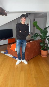 Junger Mann in einem schwarzen Sweatshirt und Jeans mit verschränkten Armen in einem hellen Wohnzimmer neben einem orangefarbenen Sofa und einer großen Topfpflanze stehend.