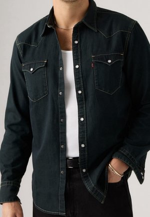 Homme portant une chemise en denim foncé boutonnée sur un débardeur blanc côtelé, avec un pantalon noir, un collier chaîne et des bracelets à un poignet.