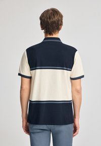 Kortärmad polo skorta med marinblå överdel och krämfärgad nederdel, med horisontella ränder i grått. Tillverkad av slät tyg med kontrasterande ribbad krage.