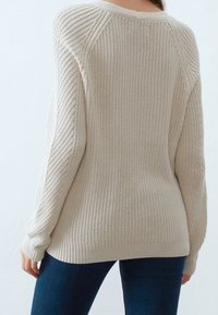 Stickad tröja i ljusbeige med ett strukturerat mönster, med en bred ribbad nederkant och raglanärmar, bärs över mörkblå jeans.