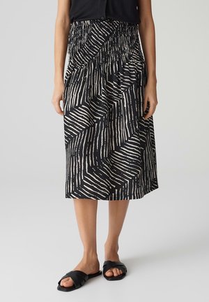 Schwarz-weiße gemusterte Midi-Rock mit geschmücktem Bund, der über vertikale und diagonale Streifen verfügt, kombiniert mit schwarzen flachen Sandalen.