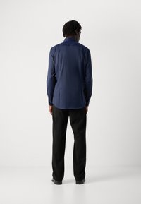 Hombre de pie de espaldas a la cámara, vistiendo una camisa de manga larga azul marino, pantalones negros y zapatos negros, contra un fondo blanco liso.
