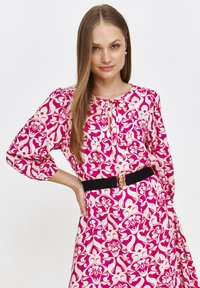 Sukienka w kwiecisty wzór w odcieniach różu i kremu, z długimi rękawami, okrągłym dekoltem oraz dopasowanym czarnym paskiem z złotą klamrą.