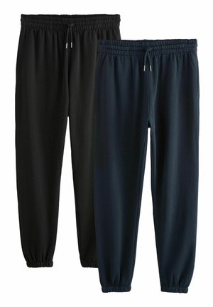 Homme portant un sweat à fermeture éclair noire jusqu'à la poitrine, pantalon de jogging noir et baskets Nike grises, debout devant un fond clair uni.