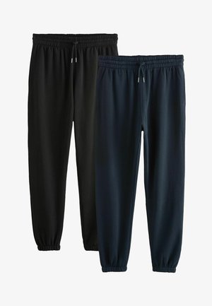 Mand iført sort kvart-zip sweatshirt, sorte joggingbukser og grå Nike sneakers, står foran en ensfarvet lys baggrund.
