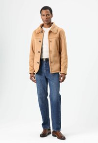 Tan faux shearling jack met kraag, vijf zwarte knopen, wit T-shirt met ronde hals, blauwe rechte jeans en bruine schoenen.