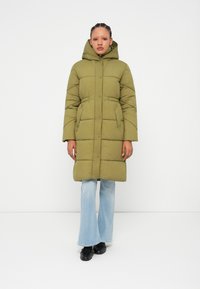Casaco puffer verde-oliva com capuz, design riscado verticalmente, duas bolsos frontais e fecho por botão de pressão, combinado com jeans claros em corte flare.