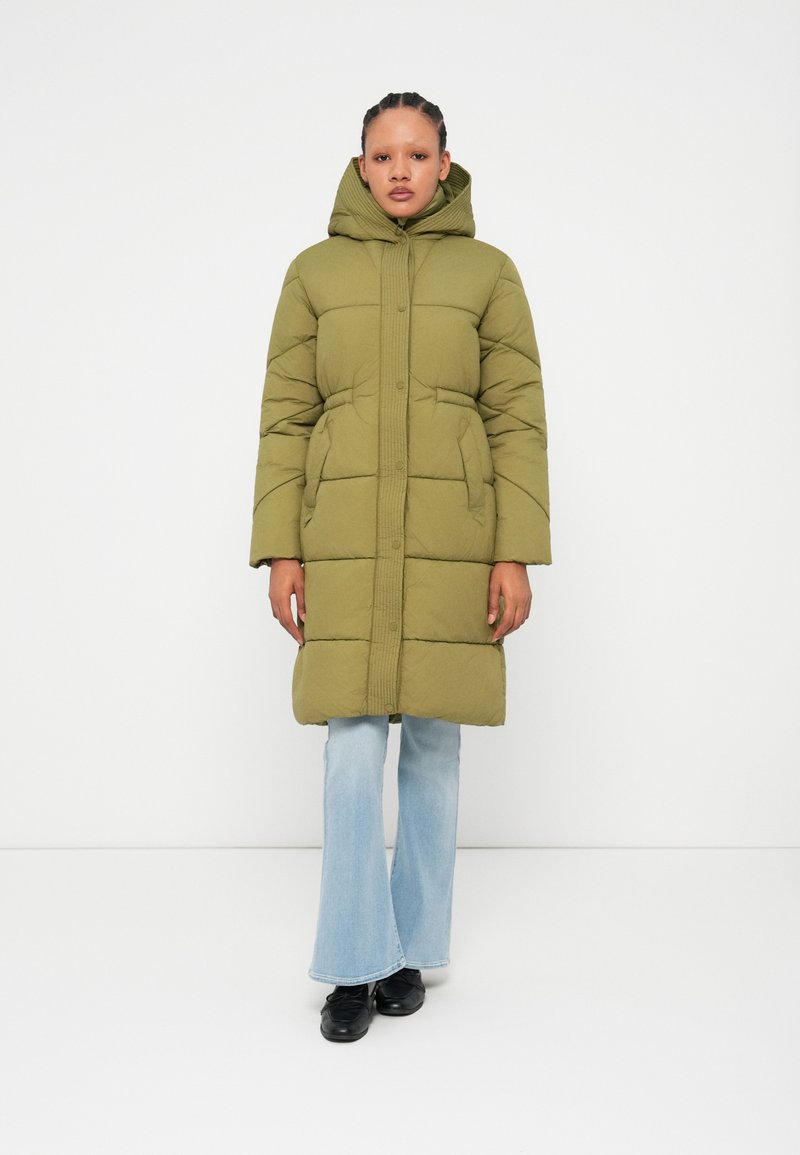 Casaco puffer verde-oliva com capuz, design riscado verticalmente, duas bolsos frontais e fecho por botão de pressão, combinado com jeans claros em corte flare.