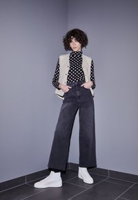 Højtaljede sorte bredbenede jeans parret med en sort og hvid hjerte-mønstret turtleneck og en lys beige quilted vest. Hvite sneakers.
