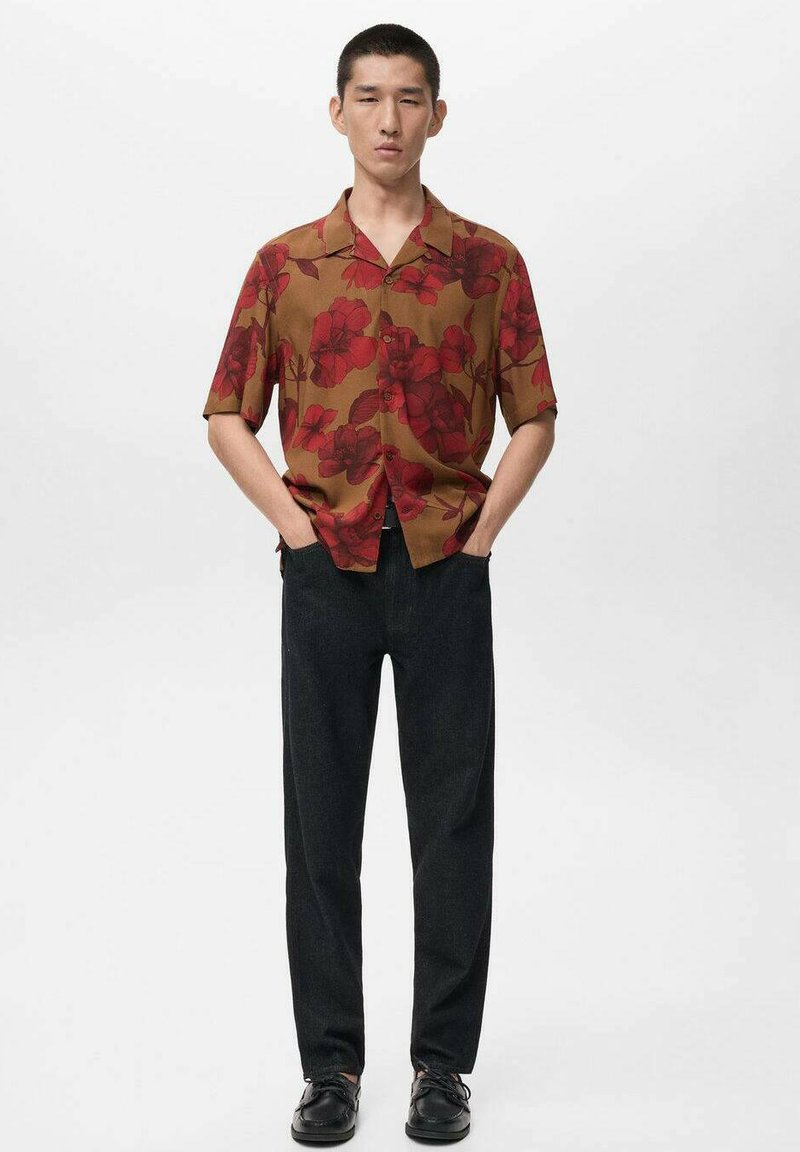 Camicia marrone a maniche corte con bottoni e motivo floreale rosso, abbinata a pantaloni scuri e scarpe nere, in piedi davanti a uno sfondo neutro.