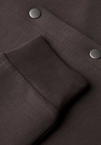 Mörkbrun tyg med en slät textur och ribbat manschett. Två metallicsnäppknappar på ytan ger detaljer.