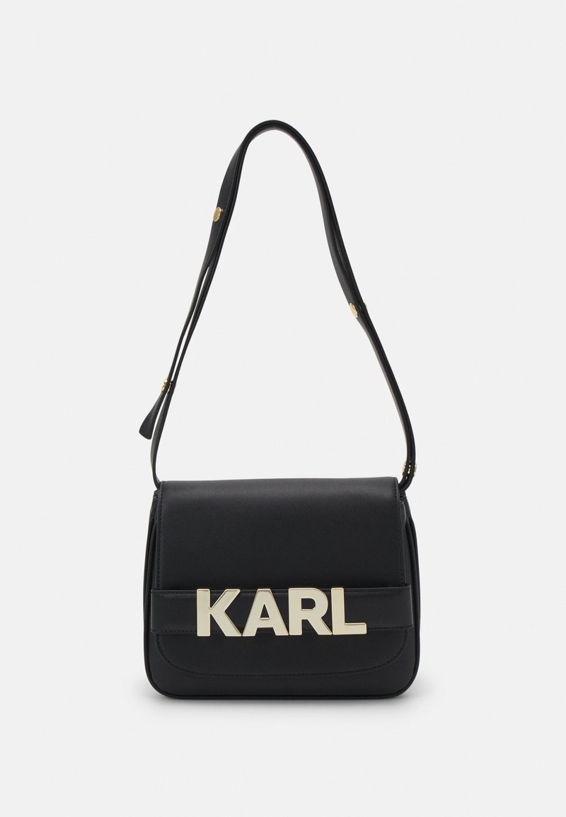 Sac à épaule en cuir noir avec un logo doré "KARL", de forme rectangulaire, fermoir à rabat plat et bandoulière ajustable. Texture lisse, design minimaliste.