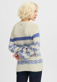 Pulover crem cu modele geometrice albastre, croială largă, mâneci raglan și tiv cu nervuri. Textură moale, lungime medie, cu etichetă pe spate.