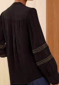 Blusa negra con mangas abullonadas, bandas horizontales decorativas de tachuelas doradas, espalda plisada y textura suave.