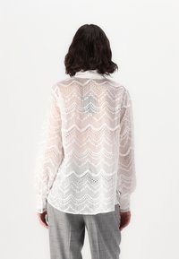 White, sheer blouse with zigzag pattern and eyelet details. Features a collar and long sleeves, paired with gray plaid trousers.