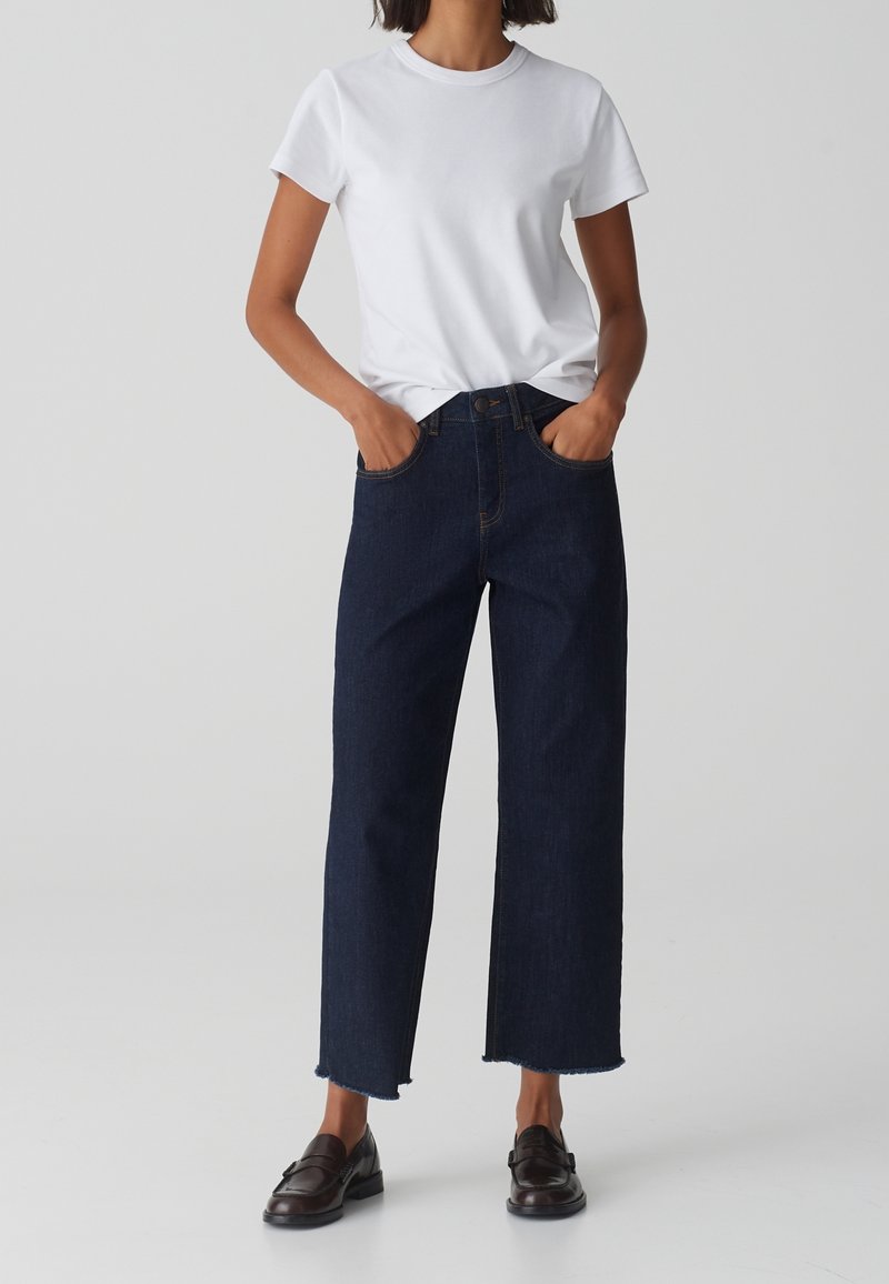 Jeans high-waisted di un blu scuro con taglio dritto, orlo grezzo e tasche frontali, abbinati a una T-shirt bianca aderente e mocassini scuri.