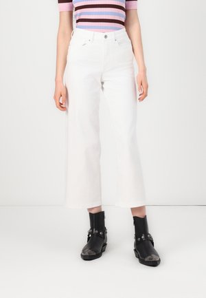 Personne portant un pantalon blanc large court, un haut à manches courtes rayé rose et marron, et des bottines noires avec des détails métalliques.