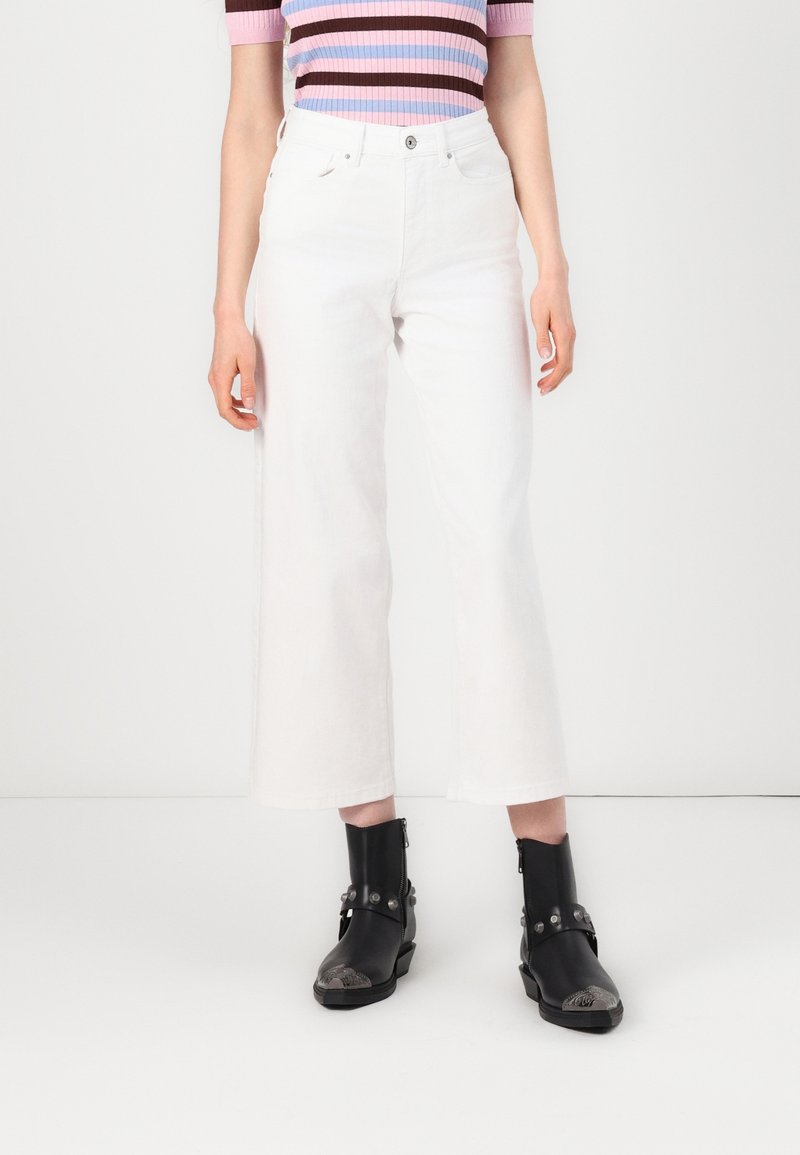 Personne portant un pantalon blanc large court, un haut à manches courtes rayé rose et marron, et des bottines noires avec des détails métalliques.