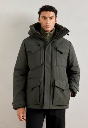 Dunkelgrüne isolierte Jacke mit Kapuze, mehreren Fronttaschen, Reißverschlüssen und einem strukturierten Stoff. Verfügt über einen hohen Kragen.