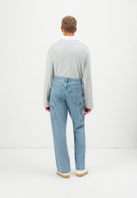 Vīrietis, ģērbies gaiši zilās vaļīgās biksēs ar kabatām un gaiši pelēkā garajās piedurknēs krekliņā, stāv, vērsts pret parastu baltu sienu.