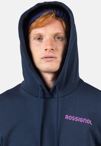 Felpa blu navy con cappuccio e coulisse, con un logo rosa "ROSSIGNOL" ben visibile sul petto. Tessuto morbido con una vestibilità comoda.