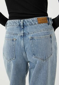 Henkilön takakuvassa vaaleansiniset farkut, joissa YAS-nahkainen merkkilappu, ja musta pitkähihaisen paita valkoista taustaa vasten.