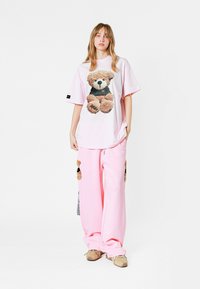 Camiseta rosa de gran tamaño con un gráfico de oso de peluche, combinada con pantalones de chándal rosas a juego que presentan acentos de oso de peluche. Diseño ligero y casual.