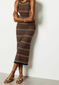 Stickad midiklänning med ett zickzackmönster i vinröd, gul och svart, i kombination med platta bruna sandaler med pärlaccenter.