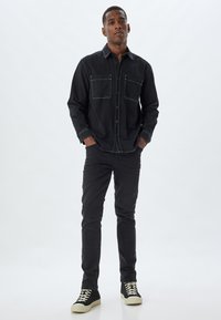 Camisa de mezclilla negra con dos bolsillos en el pecho, cierre de botones en la parte delantera y costuras en contraste. Combinada con unos vaqueros ajustados negros y zapatillas negras.