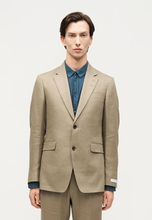 Άνδρας που φοράει μπεζ σακάκι με δύο κουμπιά και ταιριαστό παντελόνι πάνω από ένα τιρκουάζ πουκάμισο με κουμπιά, στέκεται μπροστά σε λευκό φόντο.