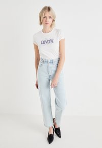 Biela tričková košeľa s krátkym rukávom a fialovým kvetinovým logom "Levi's", skombinovaná s nohavicami s vysokým pásom v svetlomodrej farbe a čiernymi špicatými topánkami.