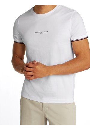 Mężczyzna w białej koszulce Tommy Hilfiger z logo na piersi i beżowych spodniach, ręce złożone przed sobą.