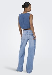 Vrouw met donker haar in een knotje modelleert een mouwloze, korte spijkerblouse en losse, lichtgewassen, hooggetailleerde jeans tegen een effen achtergrond.