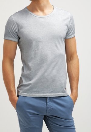 Mand iført en tætsiddende, kortærmet lysegrå t-shirt og blå bukser med hænderne i lommerne mod en ensfarvet baggrund.