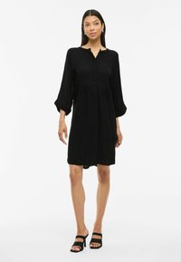 Robe noire avec un col boutonné, coupe ample, manches trois-quarts et détail de taille froncée. Tissu lisse avec un ourlet à hauteur des genoux.