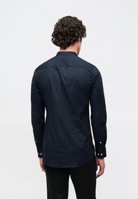 Μπλε ναυτικό πουκάμισο με μακριά μανίκια, ευθεία ρεβέρ, κουμπιά στα μανίκια και κλασικό γιακά. Λείο βαμβακερό ύφασμα με διακριτική γυαλάδα.