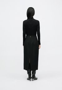 Čierny vrchný diel s dlhými rukávmi v kombinácii s dlhou sukňou s vysokým pásom, ktorá má rozparok vzadu a zlatý krúžkový detail. Jemná látka so štruktúrovaným dizajnom.