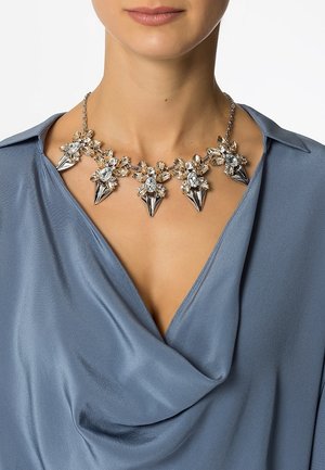 Femme portant un collier statement de couleur argent avec des cristaux clairs et ambrés ainsi que des accents métalliques pointus, sur un chemisier drapé en satin bleu.