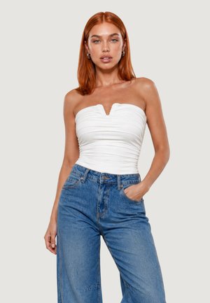 Donna con capelli rossi che indossa un top bianco senza spalline arricciato e jeans blu a vita alta, in piedi con una mano in tasca contro uno sfondo semplice.