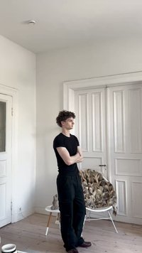 Camiseta preta, calças pretas, cabelo encaracolado; está de pé numa sala minimalista com paredes brancas, chão de madeira e uma cadeira estofada e com padrão.