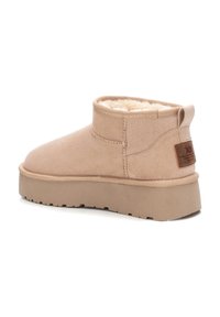 Botín de ante beige con un suave forro de piel de peluche, suela texturizada plana y un parche de cuero con marca en la parte trasera. Diseño minimalista.