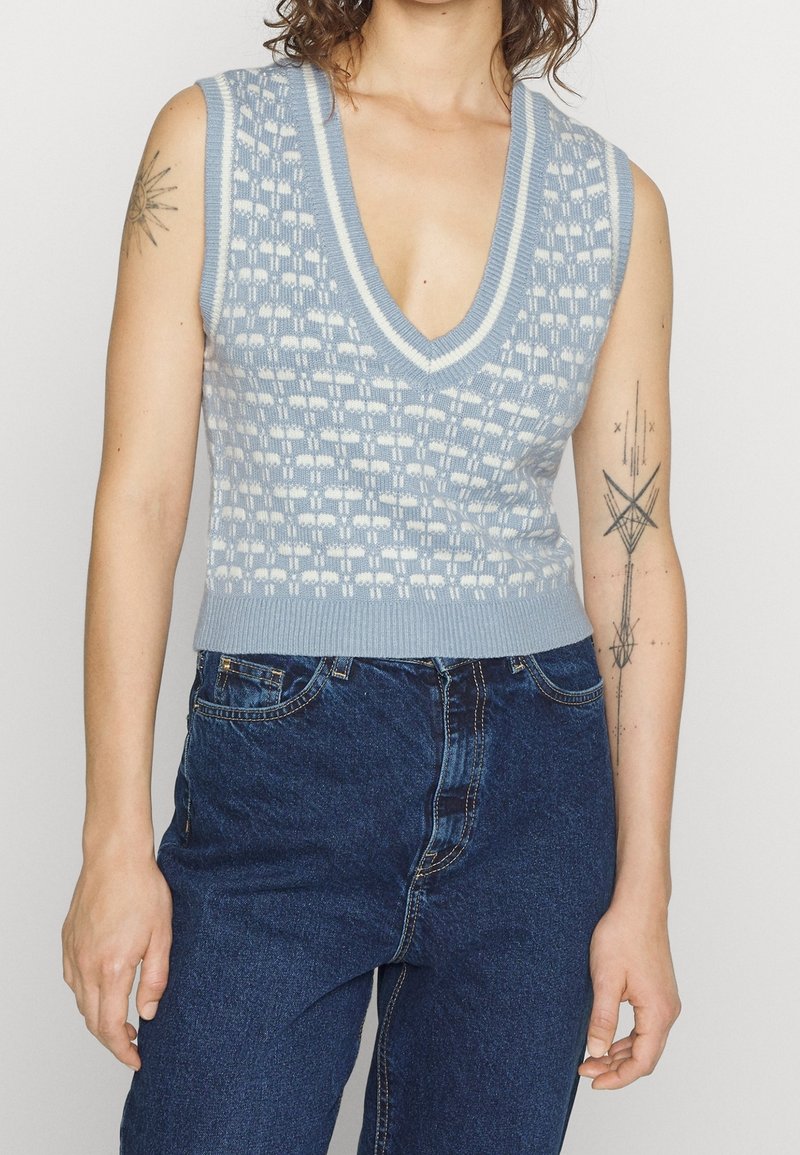 Veste tricotée sans manches à motif bleu avec un col en V profond, portée avec un jean taille haute bleu foncé, montrant des tatouages sur les deux bras.